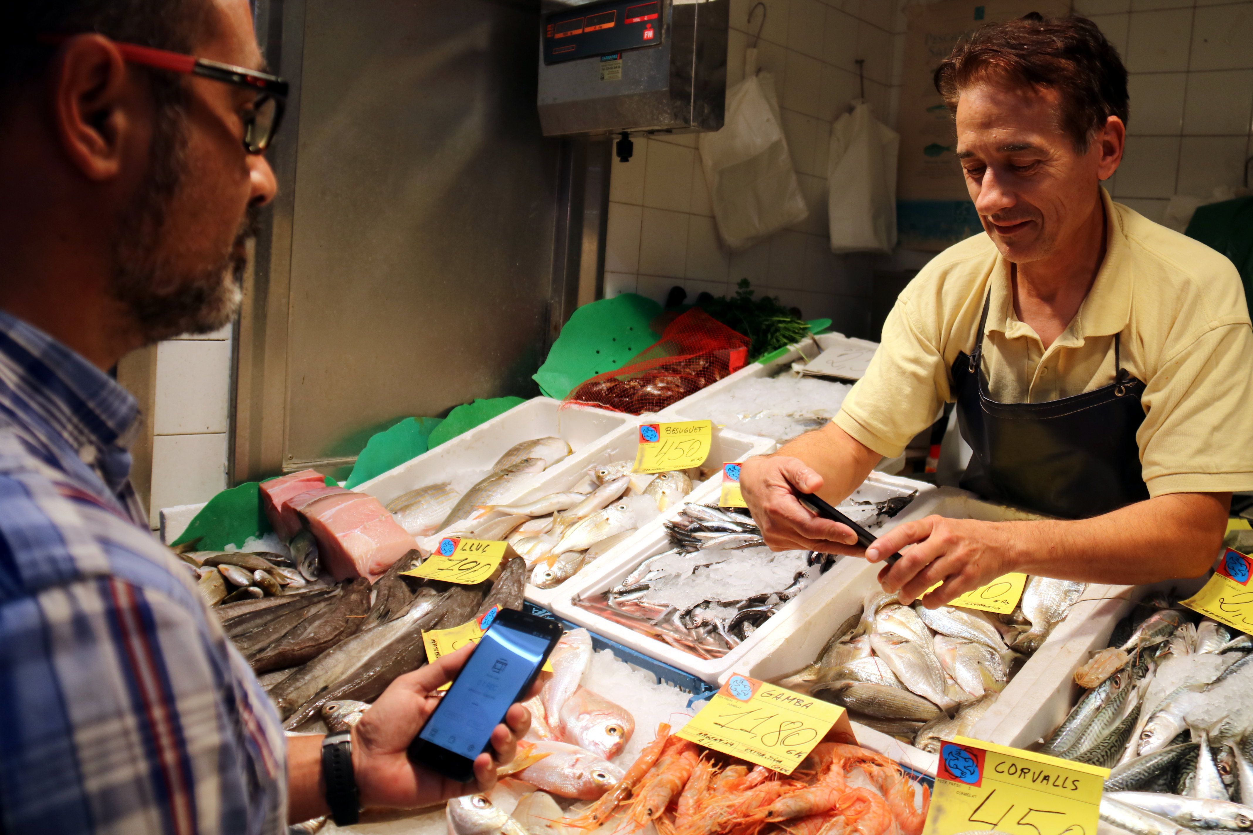 Un comerciant del Mercat del Besòs mostra l'aplicació per pagar amb la nova moneda