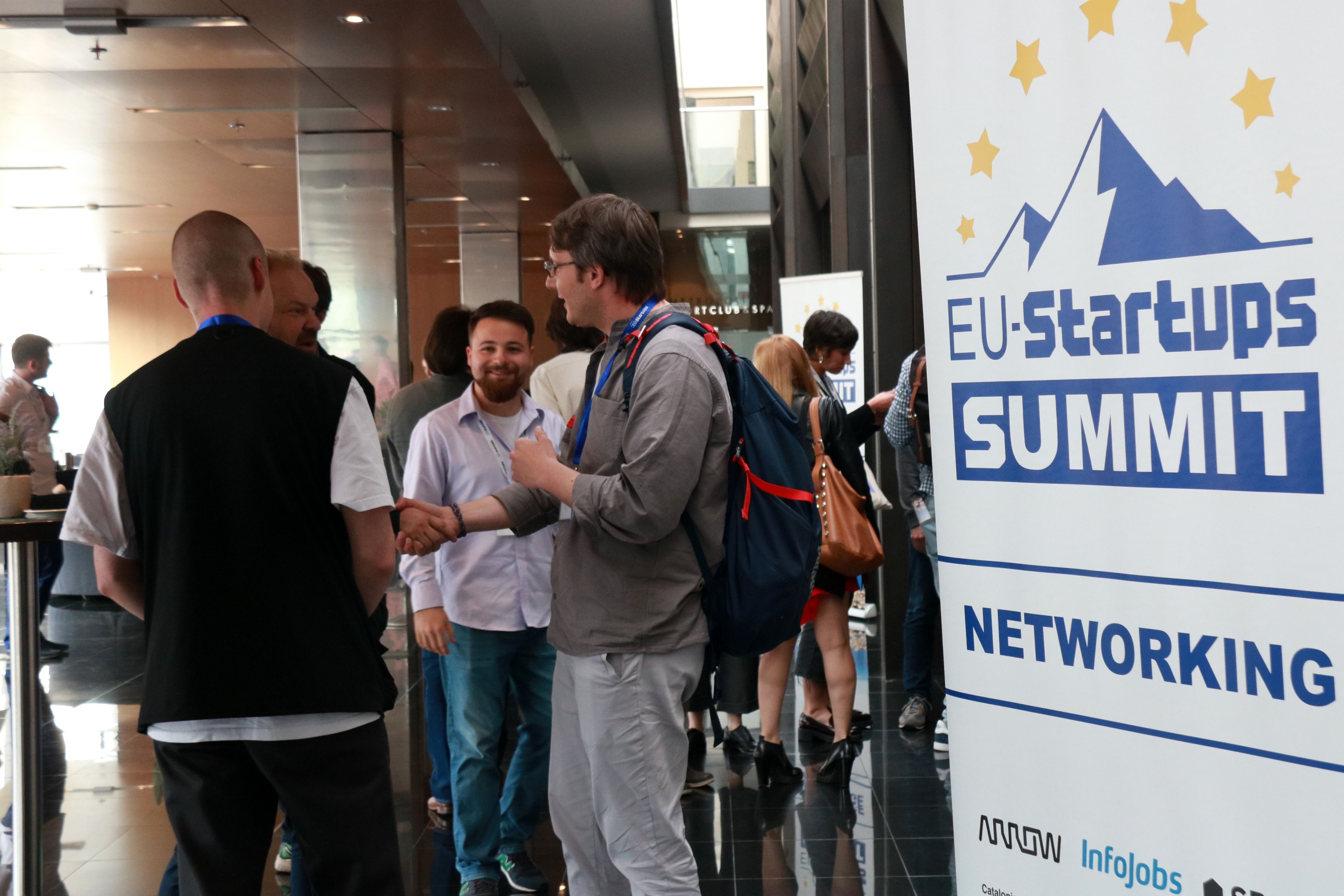 Un grup de participants a la cimera d'startups europees EU Startup Summit donant se la mà al costat d'un cartell promocional de l'esdeveniment