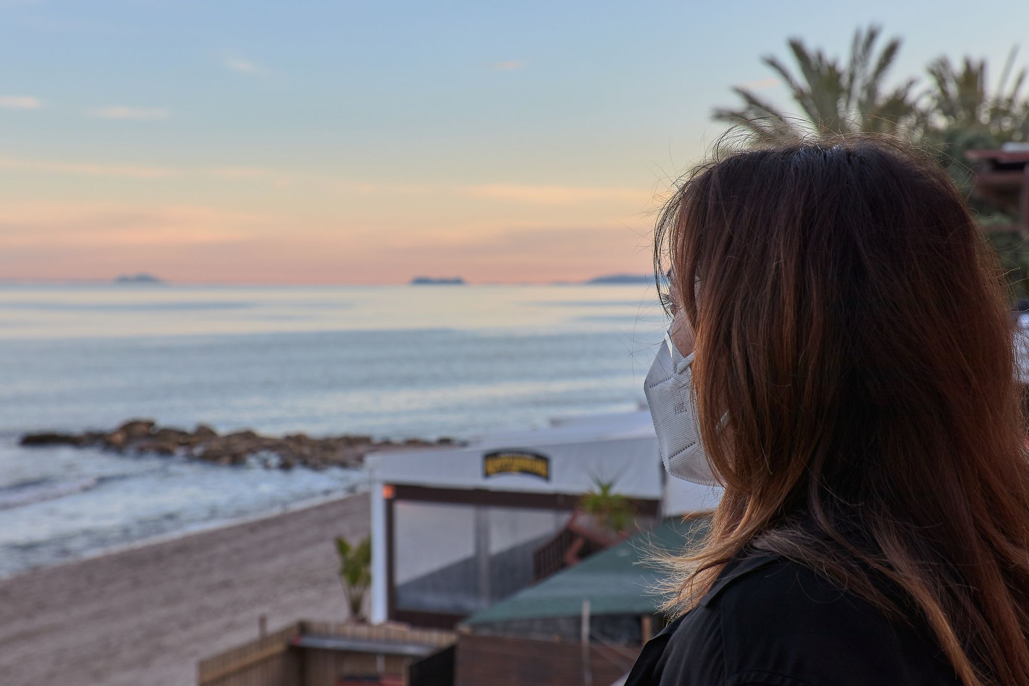 Una dona contempla una posta de sol en la platja portant mascareta | iStock