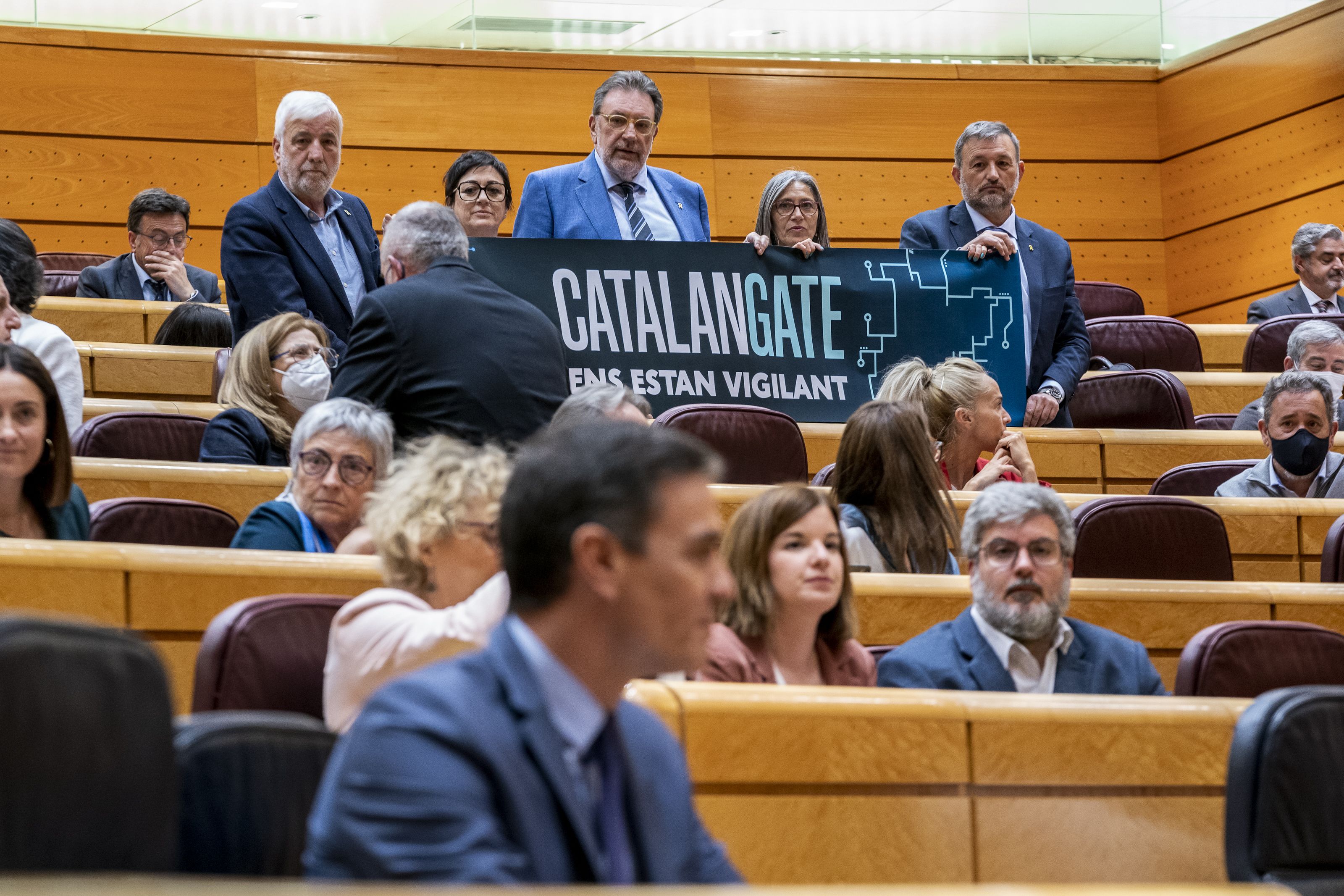 Sánchez, difuminado, con unos senadores de Junts per Catalunya (JxCat) detrás con un cartel en el que se lee: 'Catalangate ens estan vigilant' | EP