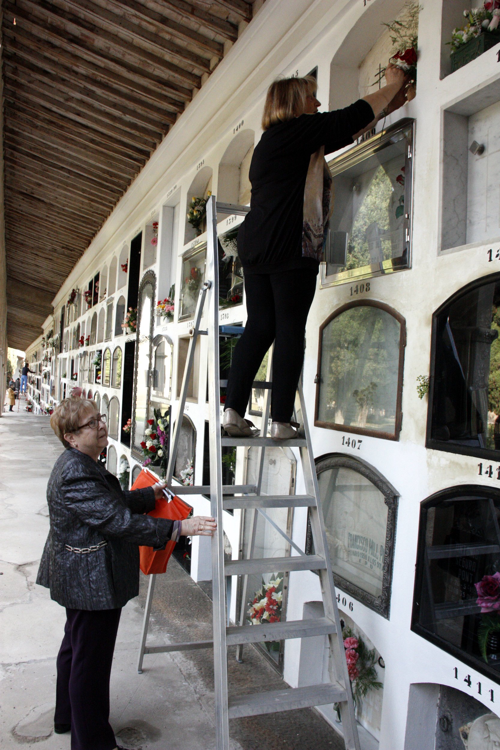 Una mujer limpia una lápida subida a una escalera al cementerio de Tàrrega.